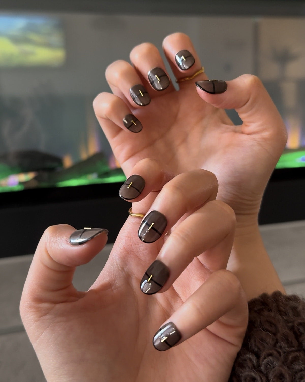 Close-up of hands wearing Smoked Alloy luxury press-on nails by Nailrative with glossy black-brown polish and gold metallic strip accents, shown in a lifestyle setting with desaturated background to highlight the modern luxury manicure.