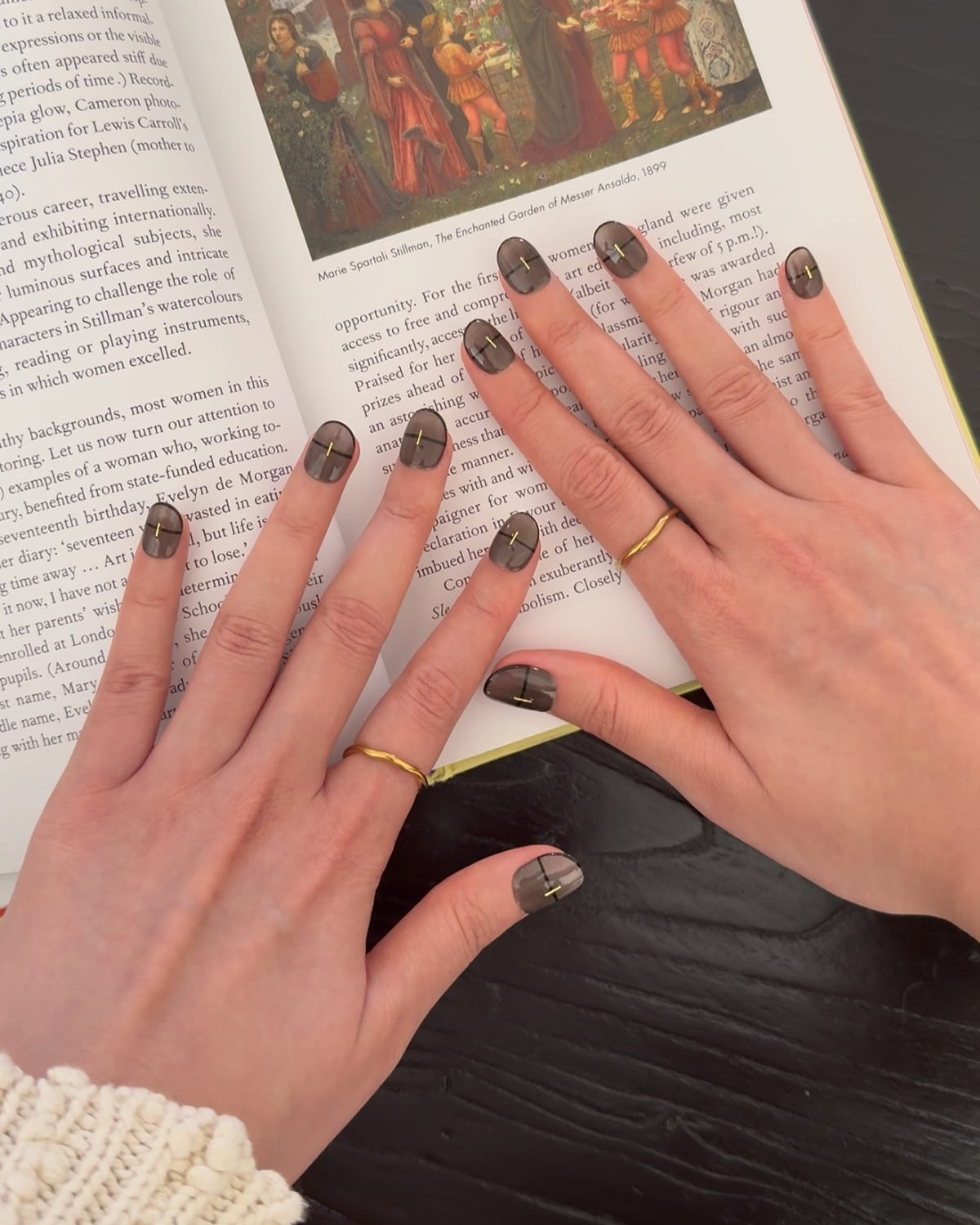 Hands wearing Smoked Alloy luxury press-on nails by Nailrative, shown resting on an open art book in soft daylight. The glossy black-brown manicure with gold strip accents contrasts subtly against a white knit sleeve, evoking quiet sophistication and modern minimalism in a lived-in lifestyle setting.