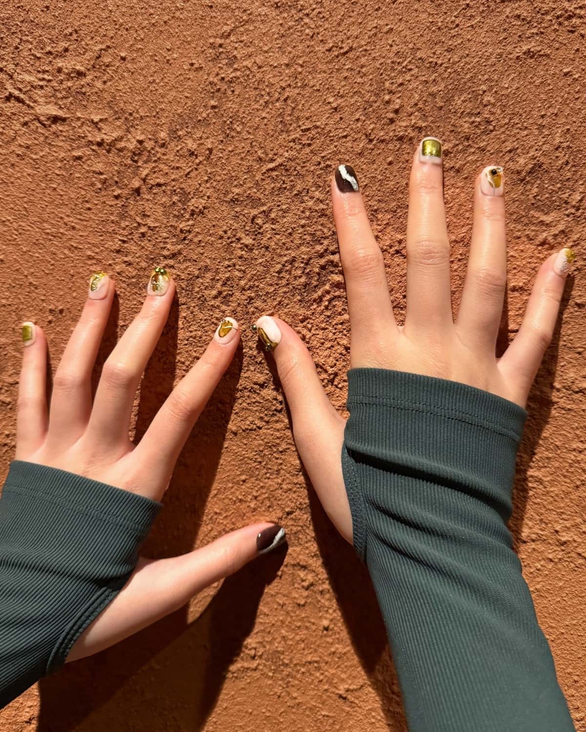 Hands with earthy luxury abstract press-on nails in green, gold, white, and brown tones, posed against a textured terracotta wall. The design features metallic accents and organic marbled patterns, inspired by natural stone and earth elements. Lifestyle manicure shot highlighting Earthen Whisper nail set by Nailrative for a modern, nature-inspired look.