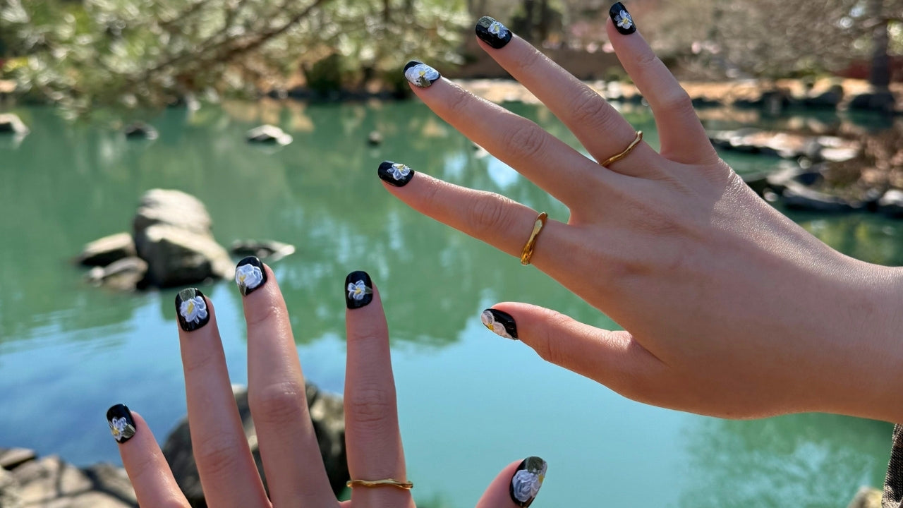 Hand wearing Nailrative’s Midnight Bloom luxury press-on nails, captured outdoors by a lake under spring blossoms — a handcrafted nail design inspired by twilight blooms and effortless femininity.