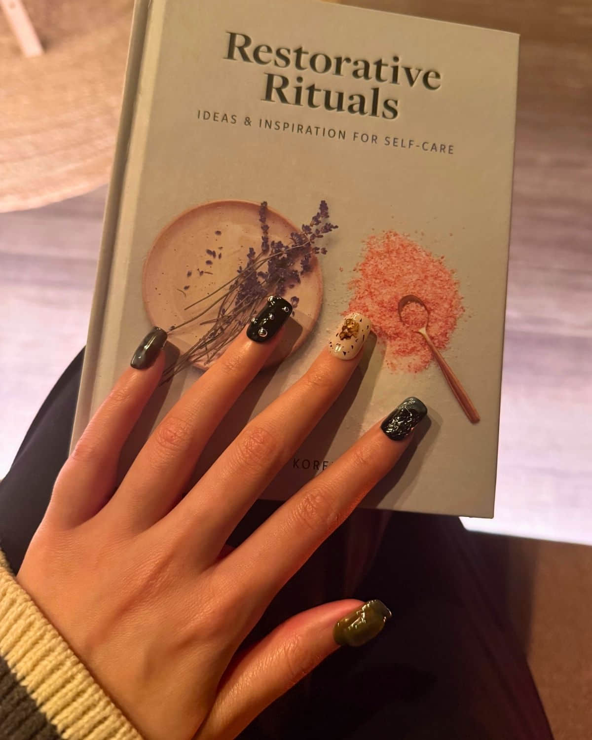 Hand with Obsidian Alchemy press-on nails holding the book Restorative Rituals, featuring black, green, and marble nail art with gold accents. Stylish manicure for modern self-care and lifestyle inspiration.