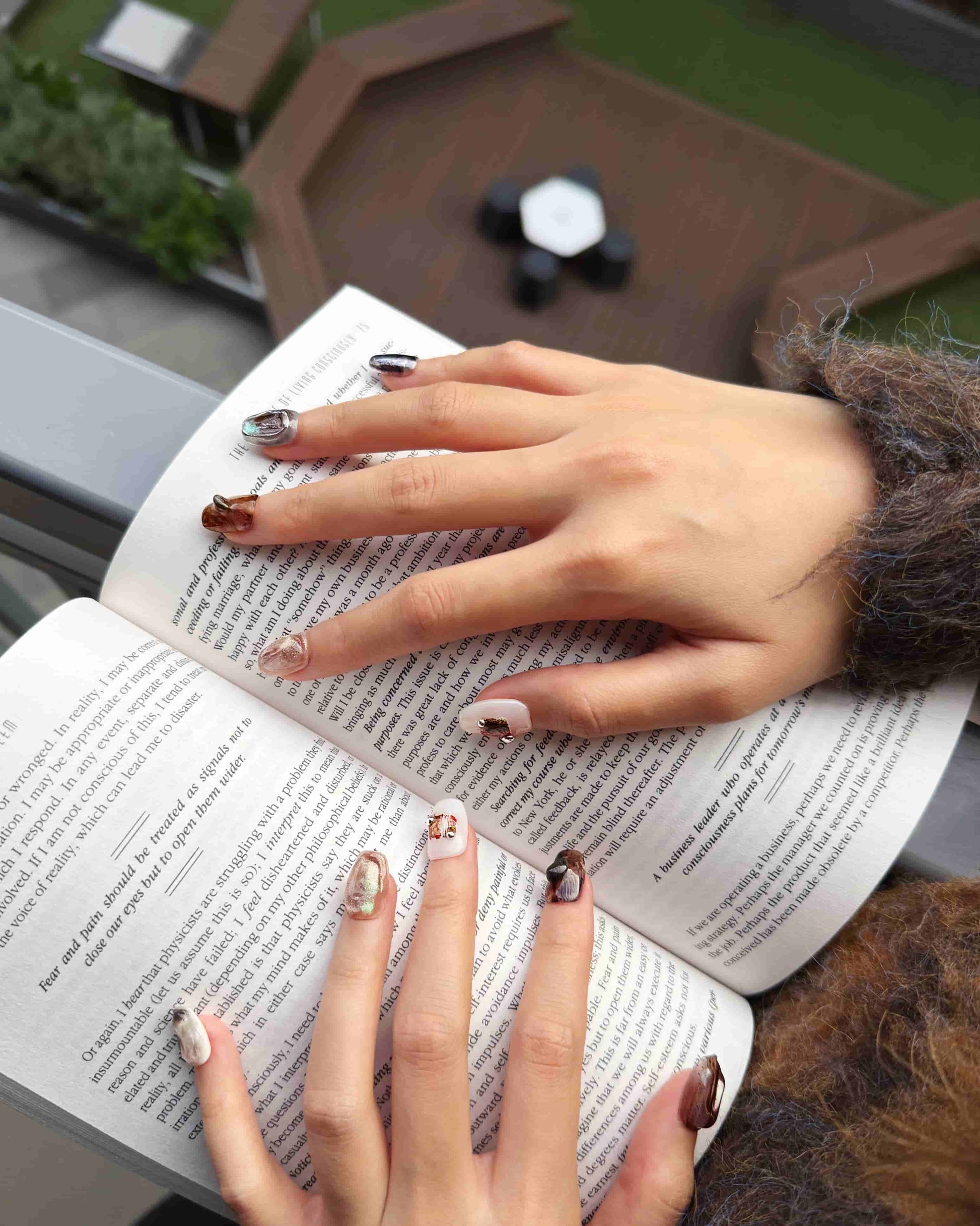 Luxury press-on nails in earthy marble tones styled while reading a book, Ethereal Terrain set by Nailrative.