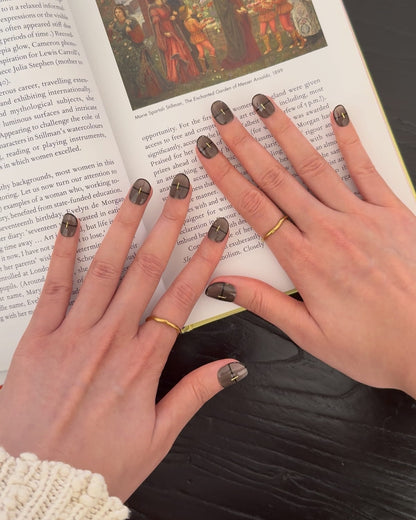 Hands wearing Smoked Alloy luxury press-on nails by Nailrative, shown resting on an open art book in soft daylight. The glossy black-brown manicure with gold strip accents contrasts subtly against a white knit sleeve, evoking quiet sophistication and modern minimalism in a lived-in lifestyle setting.