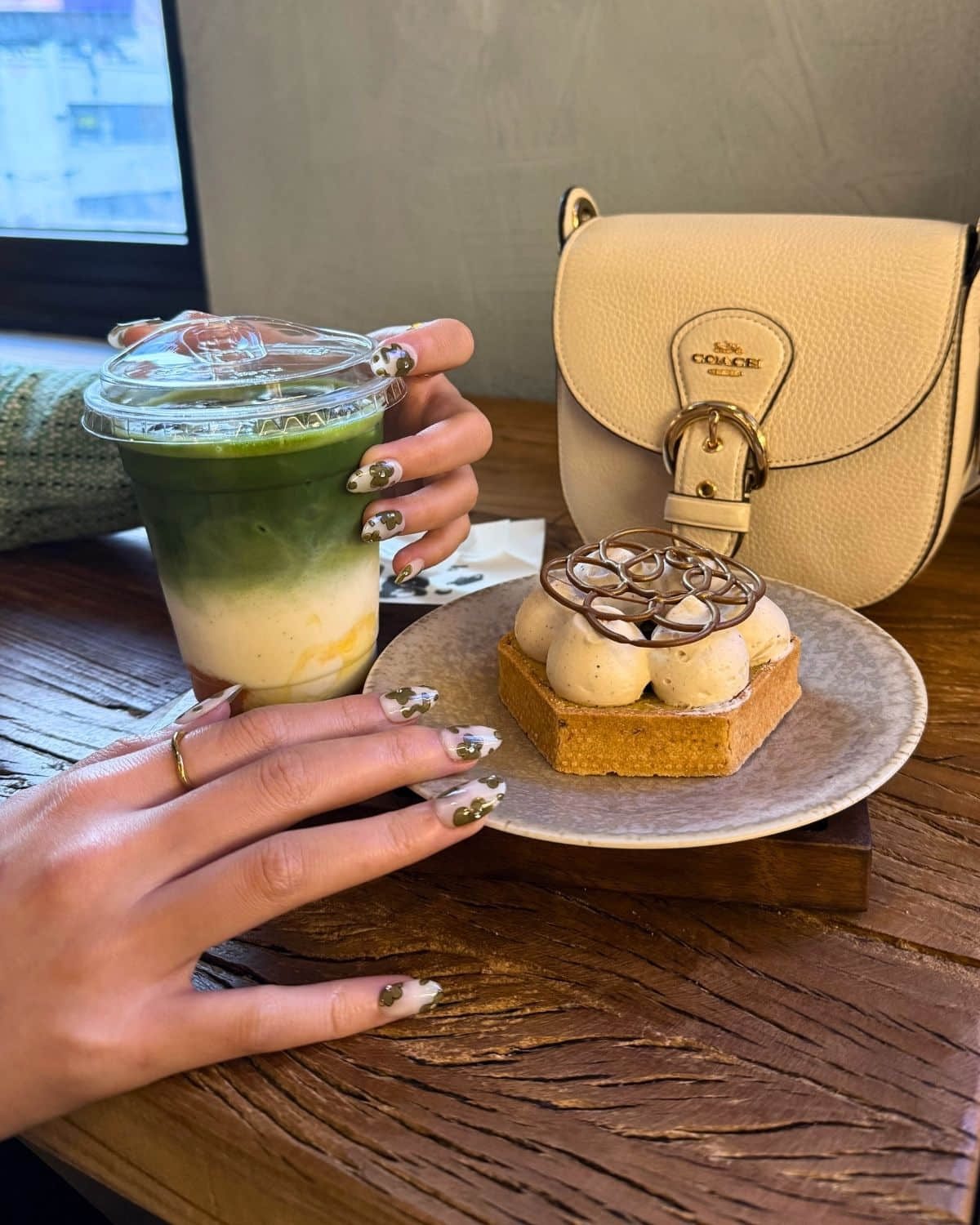 Press-on nails holding matcha latte beside cake in café.